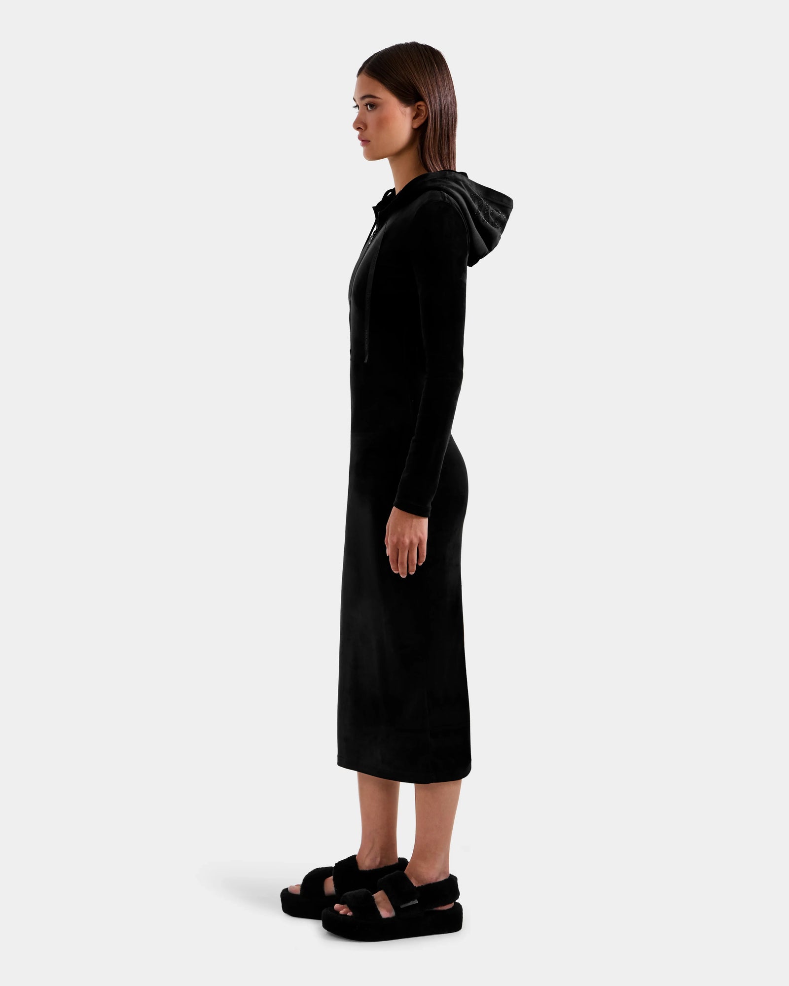 A woman stands in profile facing left, wearing the Rudsak ISABELLA - L525502 Black velour hooded midi dress with long sleeves, paired with black platform sandals against a plain light background.