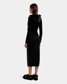 A woman with straight brown hair stands with her back to the camera, wearing Rudsak’s ISABELLA - L525502 Black velour hooded midi dress and black platform shoes against a plain light background.