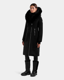 A woman poses facing forward against a plain light background, wearing the Rudsak JANIS - L125558 Black winter coat with a large faux fur hood, full front zipper, and zippered pockets, paired with black leggings and black boots.