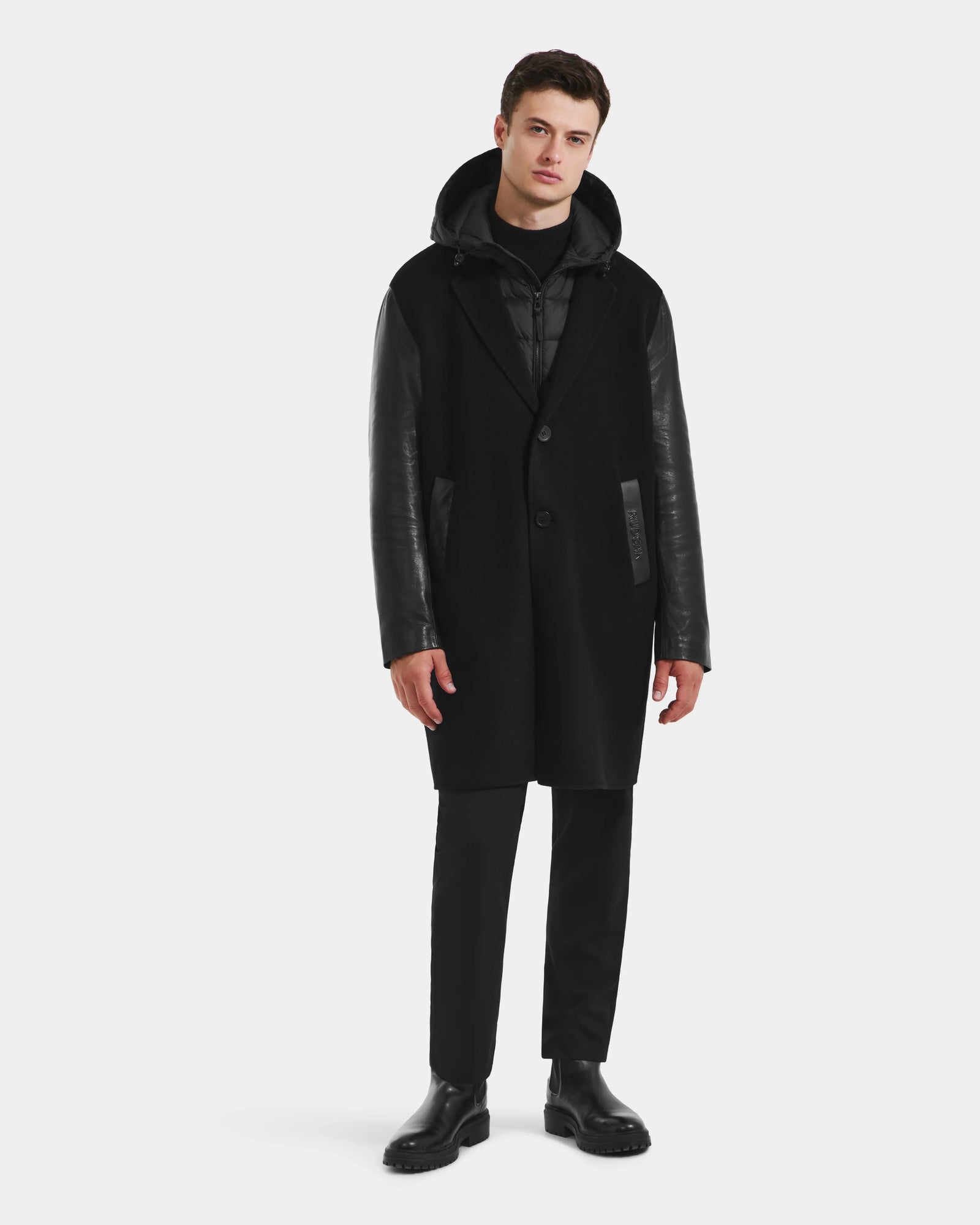 A man stands facing forward against a plain white background, wearing the Rudsak JESSE - M125504 Black men's wool coat with leather sleeves over a black hoodie, matched with black pants and shoes.