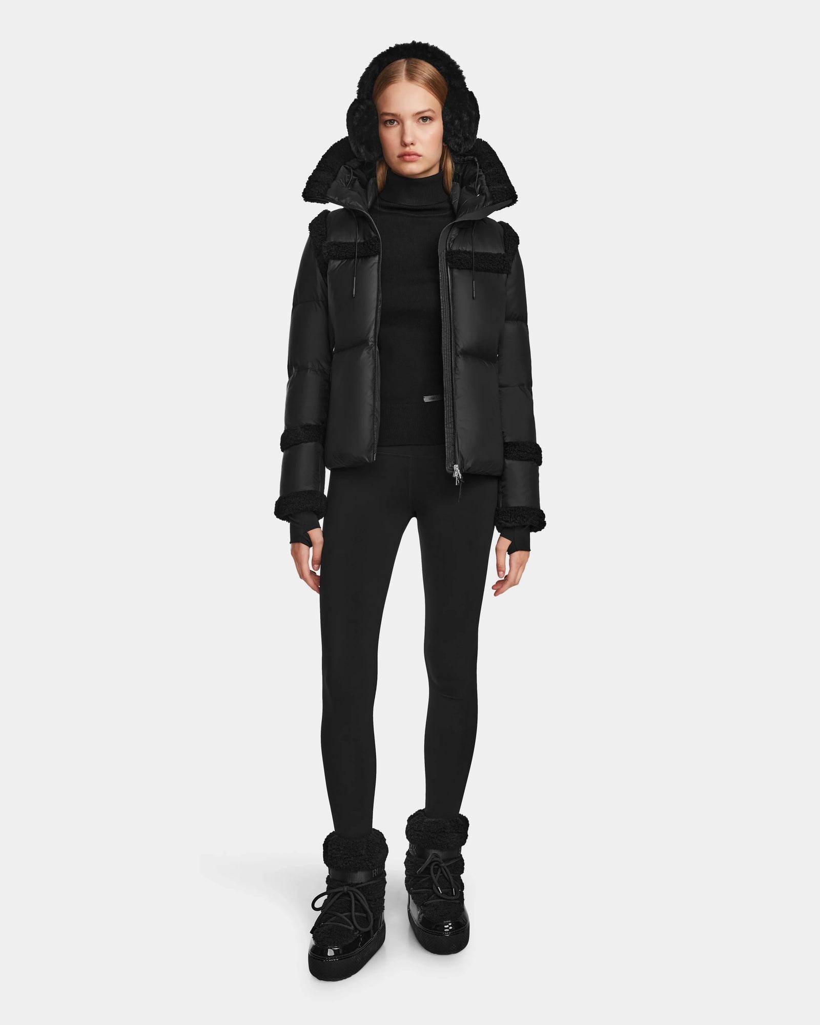 A woman faces forward, wearing the Rudsak JULIANA - L124509 Black/Black short down puffer jacket with a fur-lined hood, paired with a black turtleneck, leggings, and boots against a plain light background.