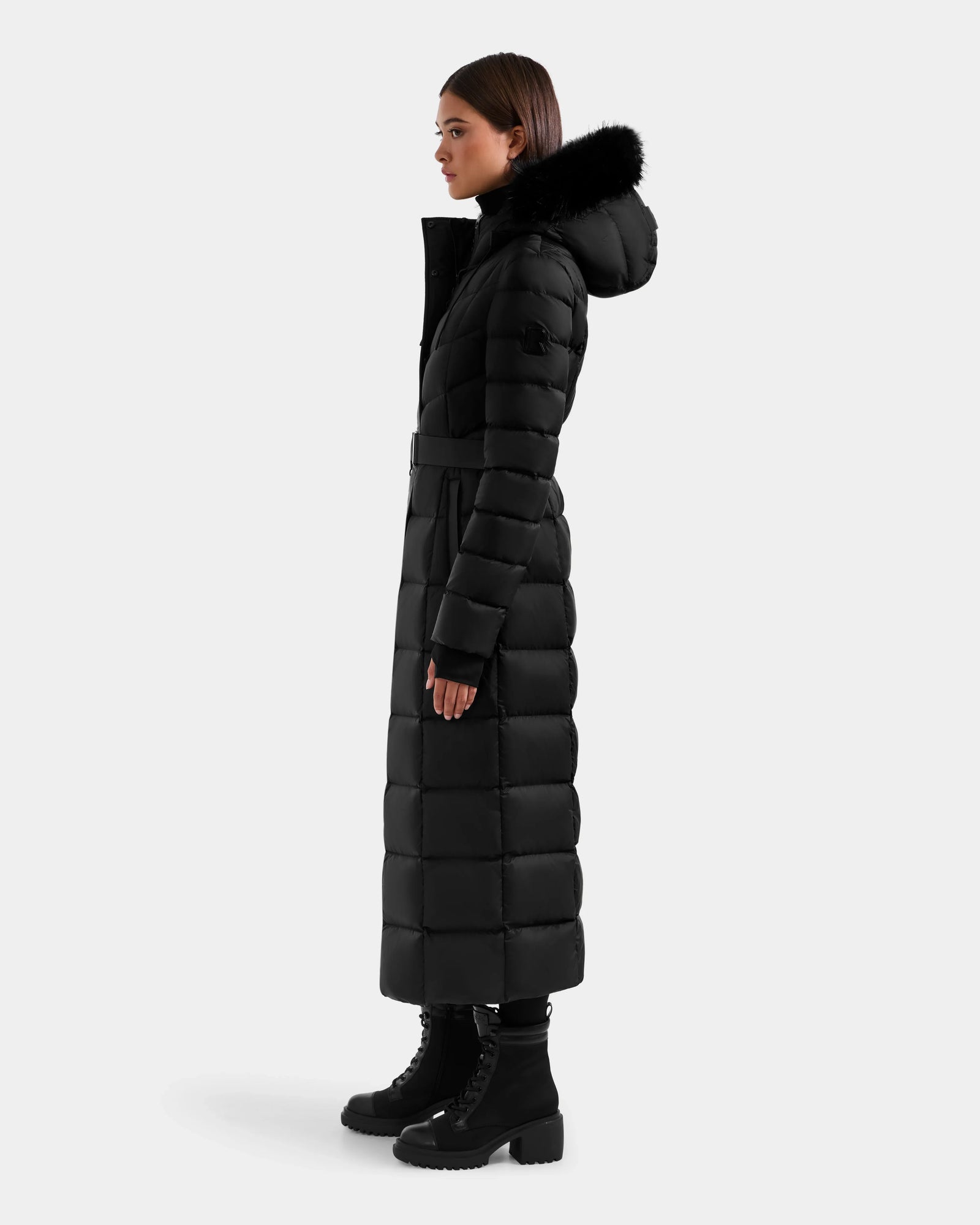 A woman stands in profile against a light grey background, wearing the Rudsak JULIETA M-L - L125559 Black maxi down puffer with a belted waist, black fur-trimmed hood, and black lace-up boots.