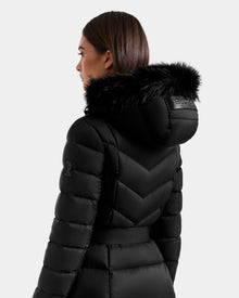 A woman with straight brown hair models the Rudsak JULIETA M-L - L125559 Black coat, a quilted maxi down puffer with faux fur-trimmed hood and weather-resistant shell, shown from the back and side on a light gray backdrop.
