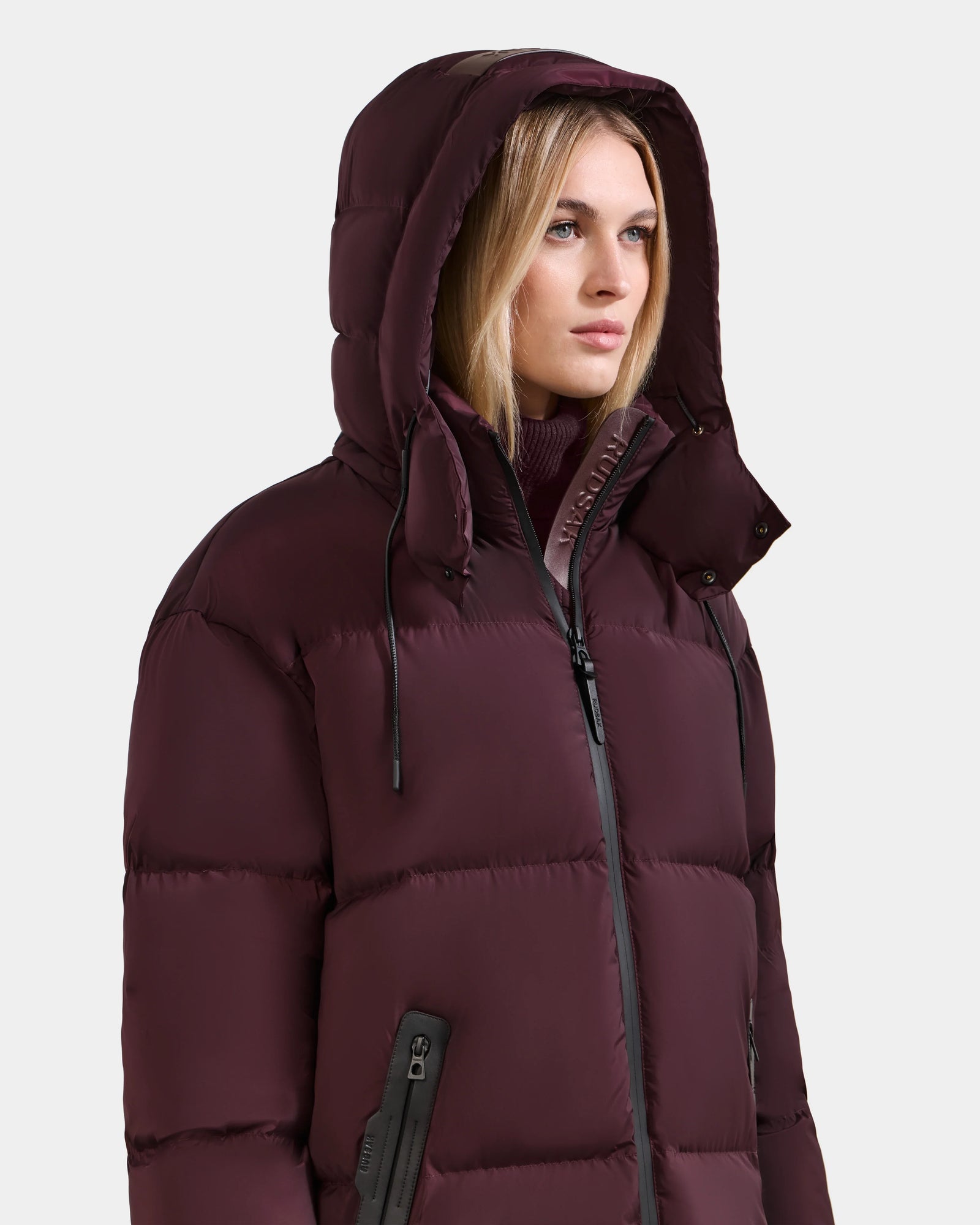 A woman models the Rudsak KARLEE L - 8125682 Bordeaux, a long maroon down puffer with hood, front zipper, and zippered pockets. She stands against a plain light background, gazing slightly to the side with a neutral expression.