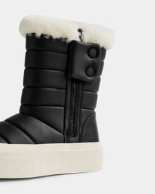 A close-up of Rudsak’s KATHIE - L225516 Black women’s mid boot, featuring black quilting, a side zipper, two buttons, white faux fur lining, and a thick white textured rubber sole.
