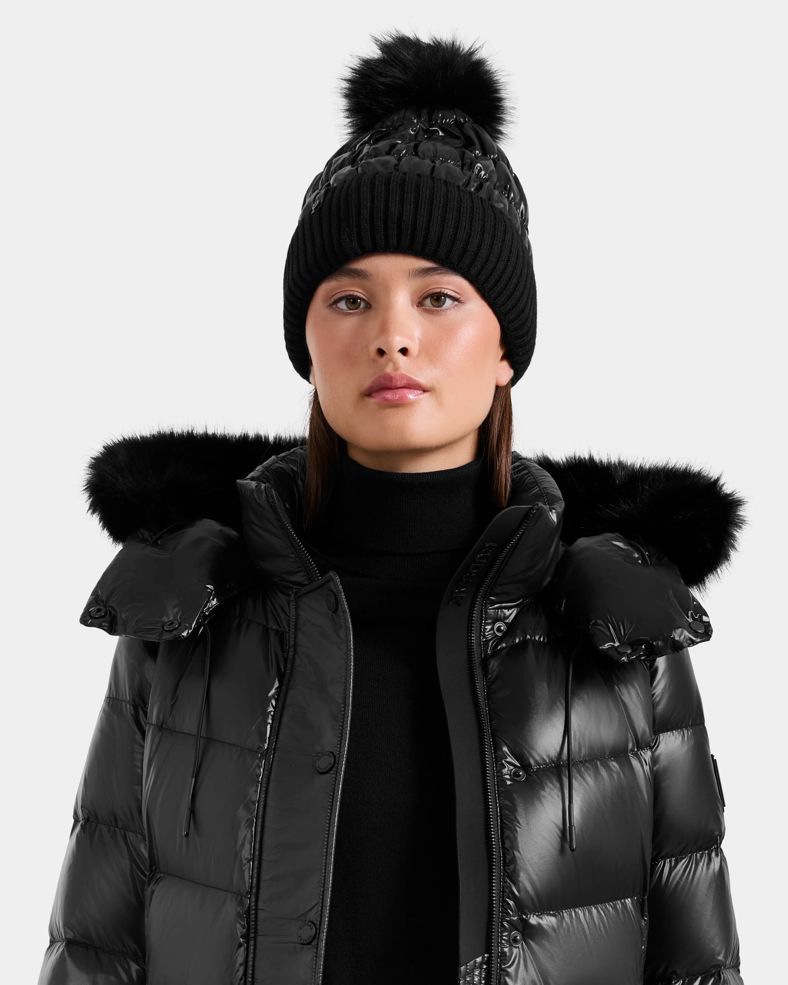 A woman in the Rudsak LANYA - L423514 Metallic Black puffer jacket with a fur-trimmed hood and a women's pom pom hat stands facing forward against a plain light background.