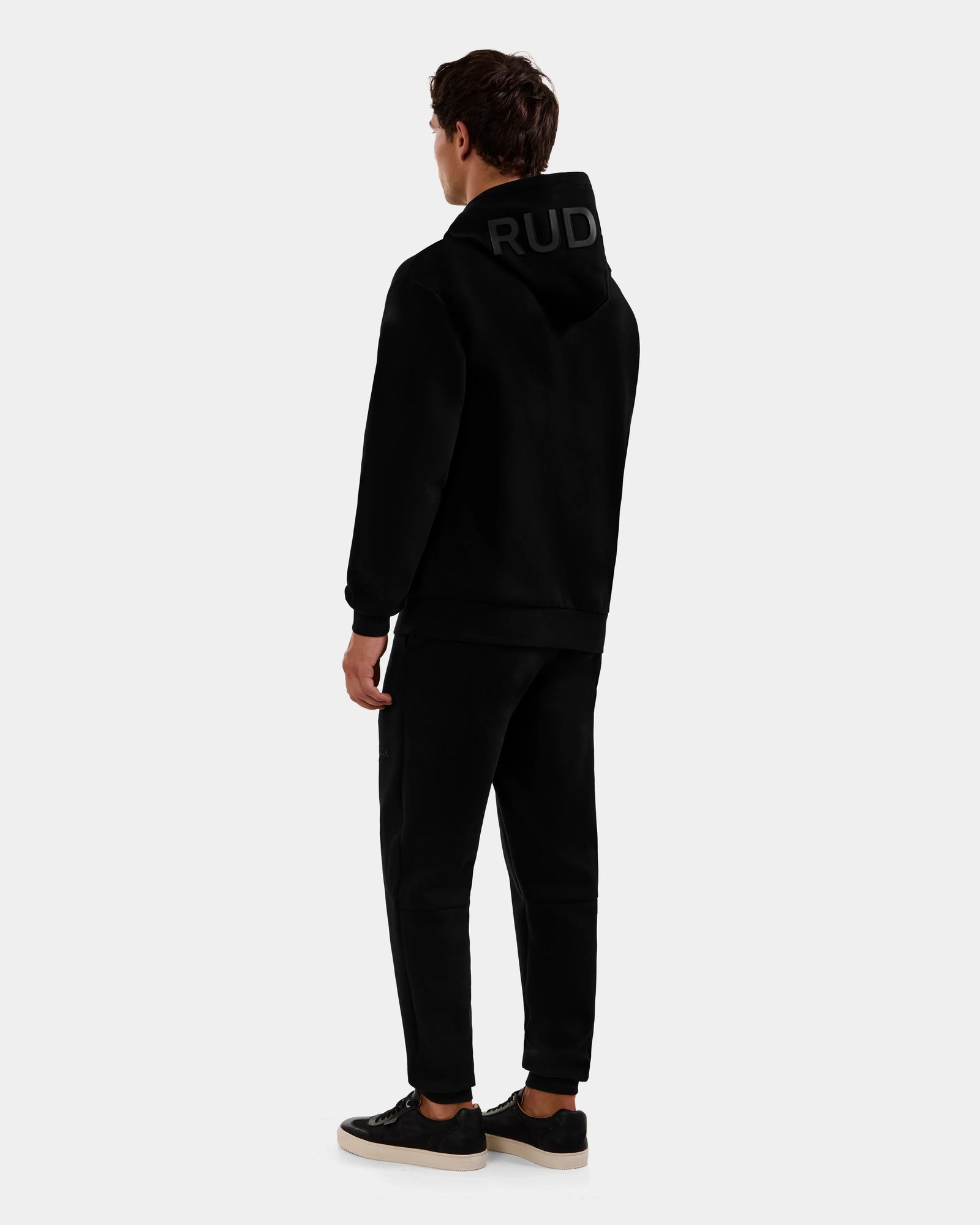 A person stands facing away, wearing the Rudsak LEROY - U525509 Black M cotton-blend unisex hoodie with "RUDI" text on the hood, black pants, and black sneakers with white soles against a light gray background.