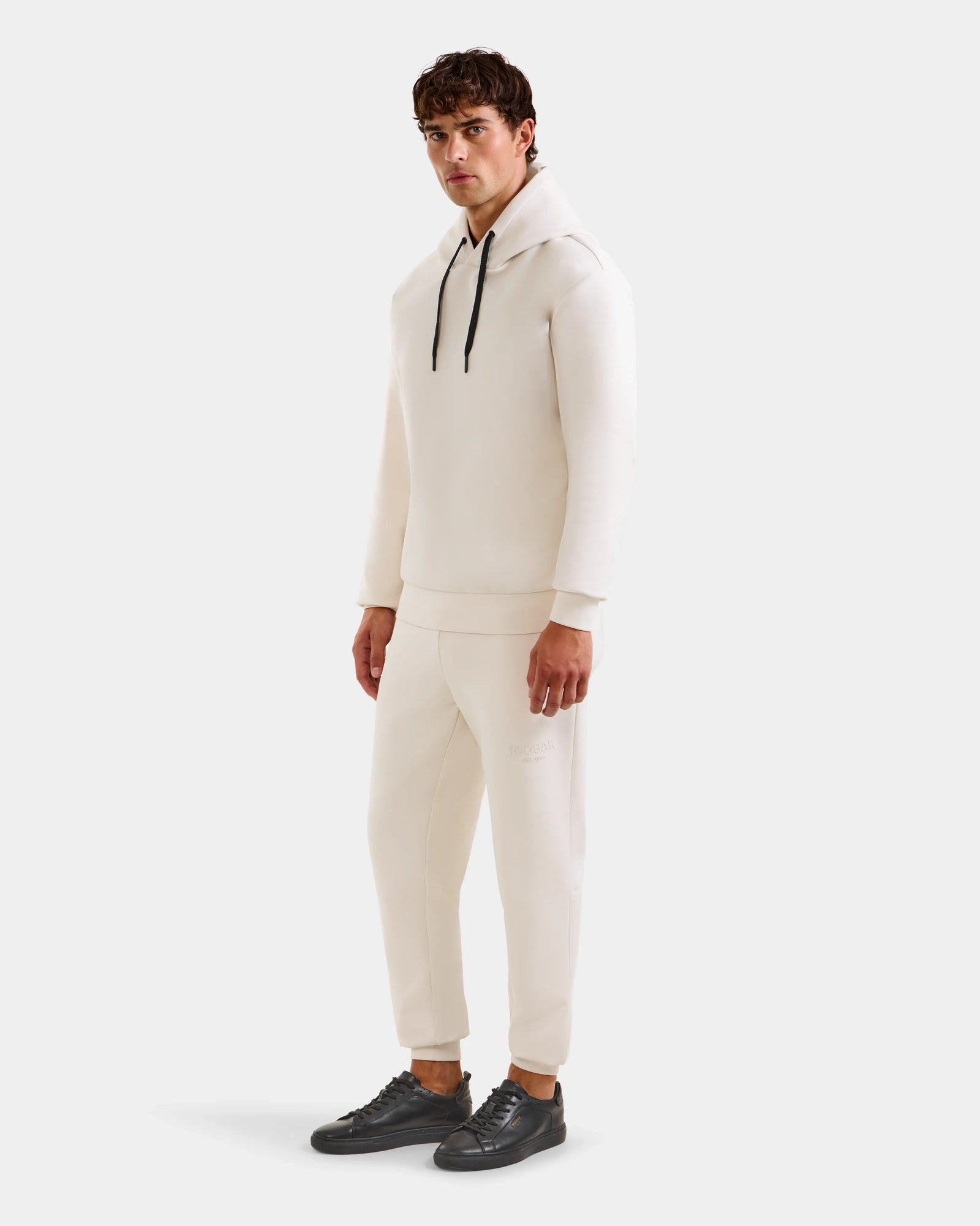 A man with short brown hair wears the Rudsak LEROY - U525509 Vanilla M premium off-white hoodie and matching joggers, paired with black sneakers, standing simply styled against a plain light background.