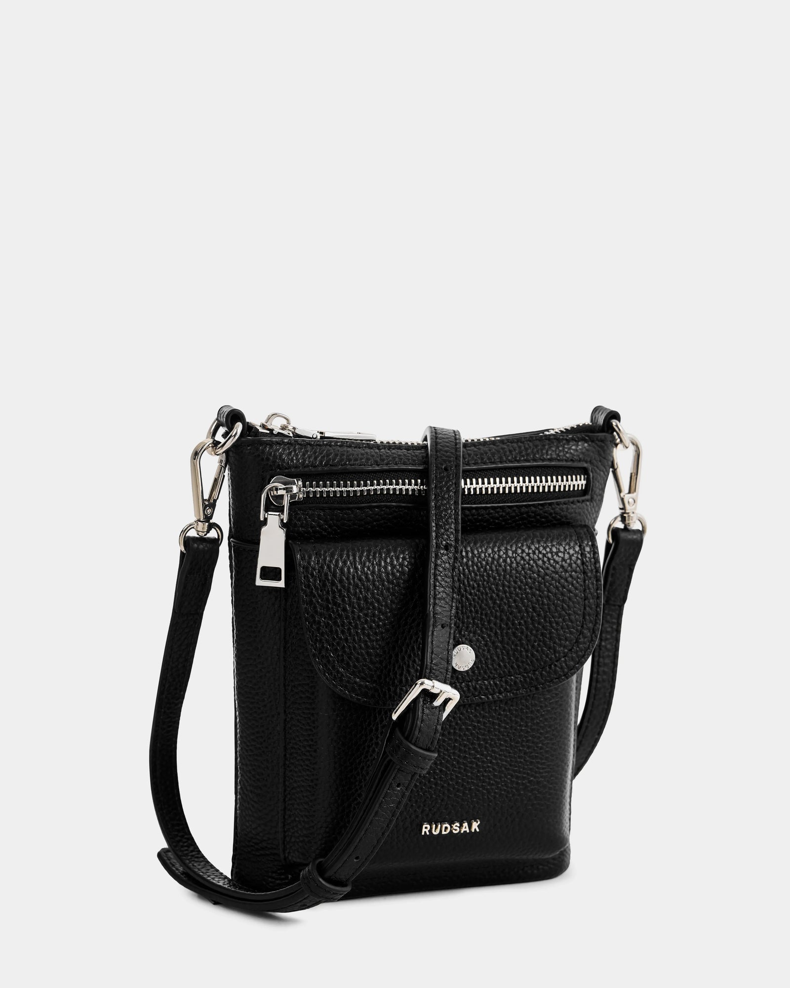 The Rudsak LILA - L325513 Black is a small women's crossbody bag in pebbled leather with silver hardware, a front flap pocket, top zipper closure, adjustable strap, and the RUDSAK logo in silver on the front.