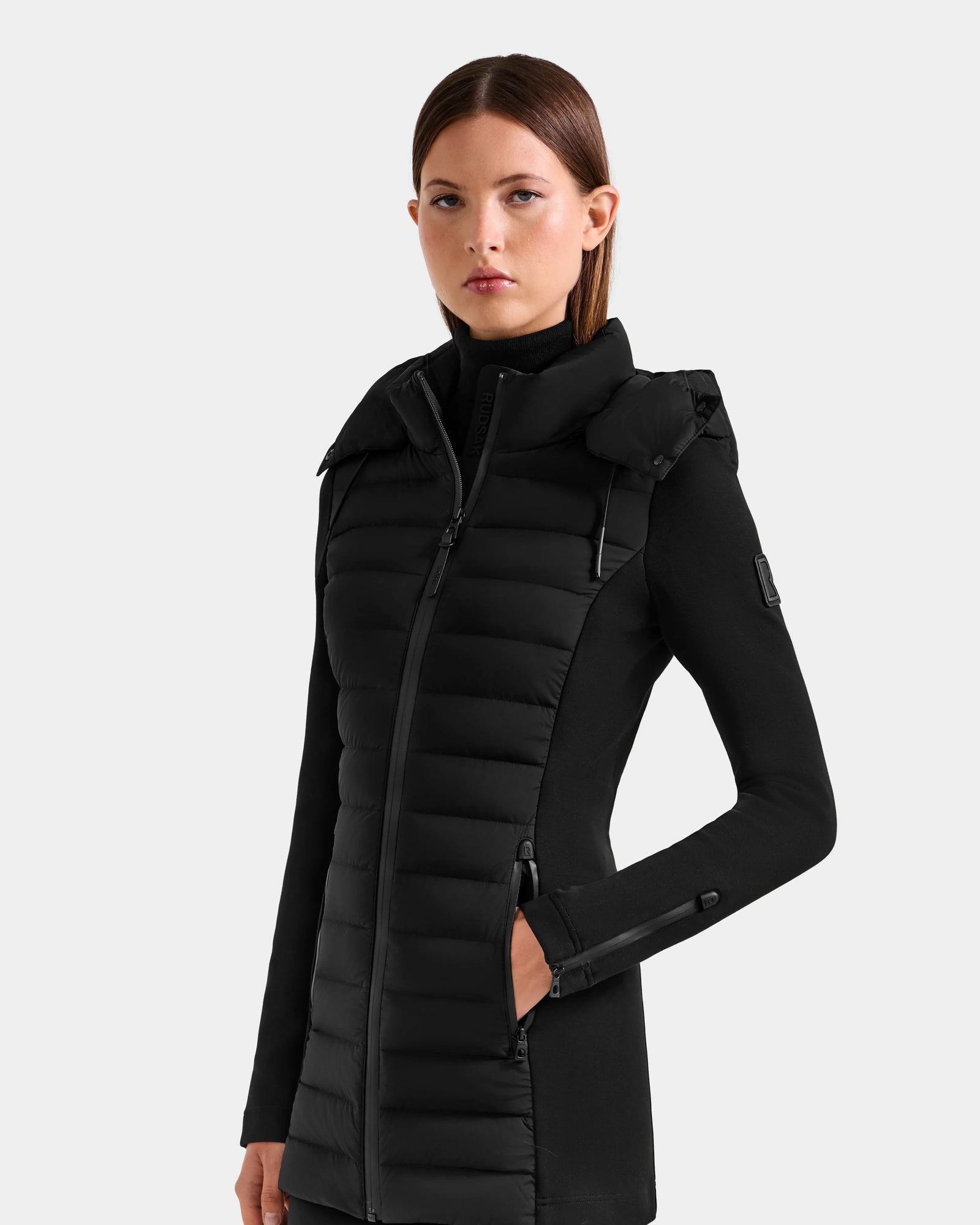 A woman wearing the Rudsak MARRY M - L125519 Black women’s mid-length hybrid coat with a high collar and zippered pockets stands against a plain light gray background, looking at the camera with a neutral expression.