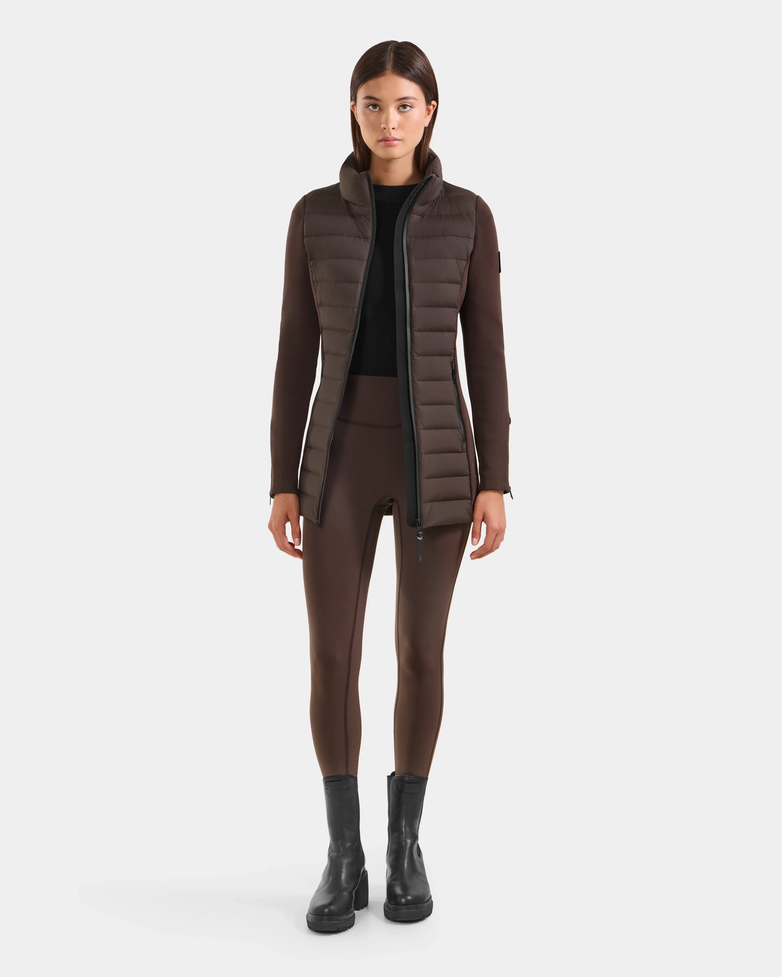 A woman faces forward wearing the Rudsak MARRY M - L125519 Espresso hybrid jacket with down insulation, paired with matching brown leggings, a black top, and black ankle boots against a plain white background.