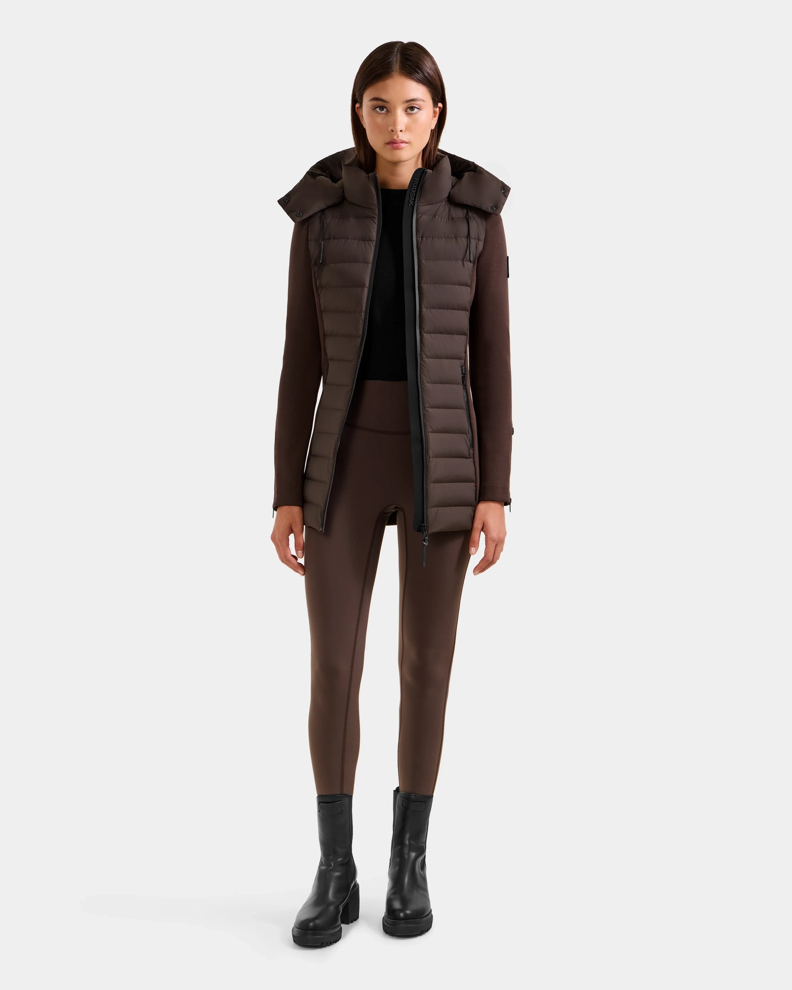 A woman stands against a plain background wearing the Rudsak MARRY M - L125519 Espresso women’s hybrid jacket, brown leggings, a black top, and black ankle boots. She looks straight ahead with a neutral expression.