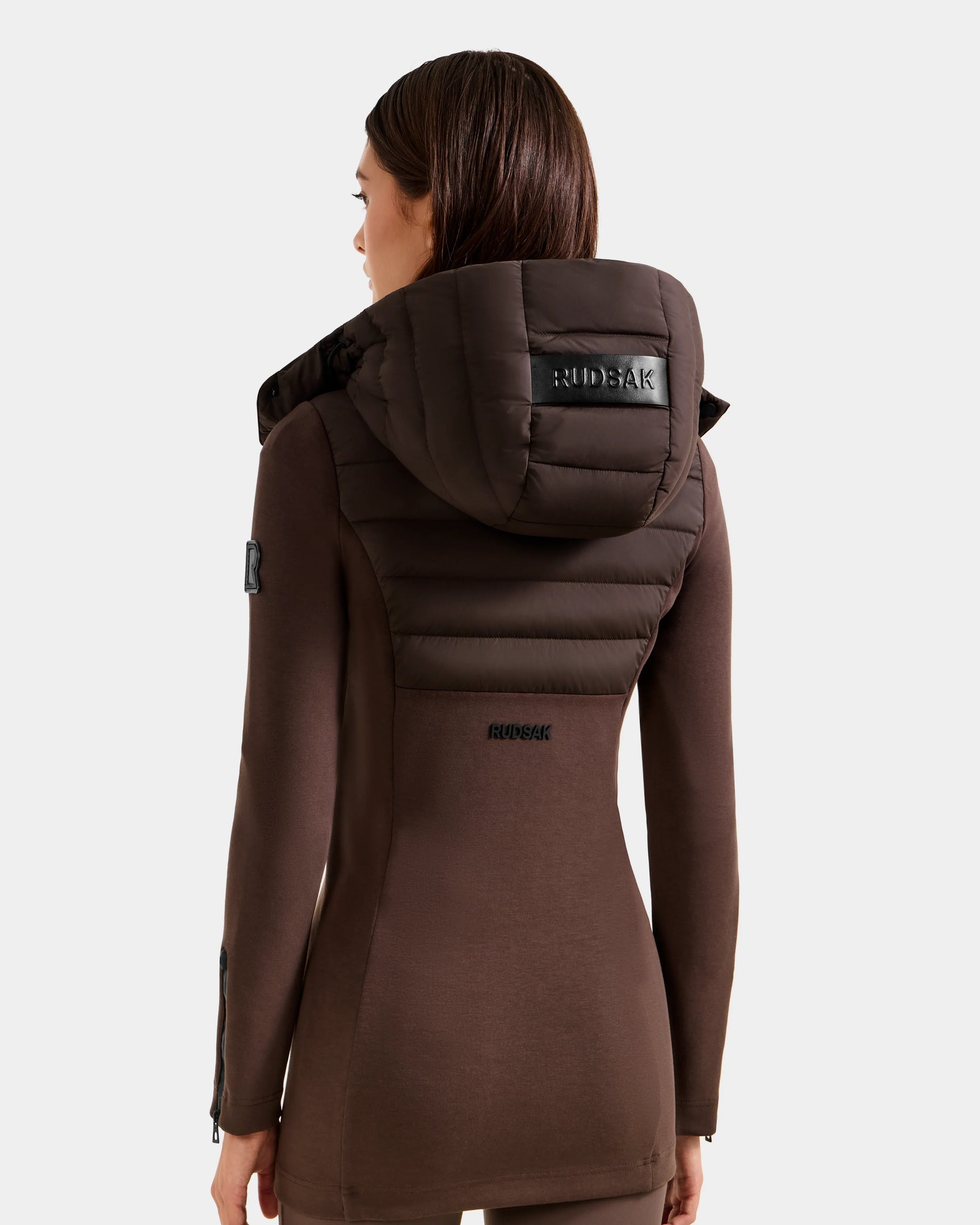 A woman with straight brown hair is seen from behind wearing the Rudsak MARRY M - L125519 Espresso long, fitted women’s hybrid jacket with a padded hood. The black Rudsak logo accents the hood and upper back against a plain light background.