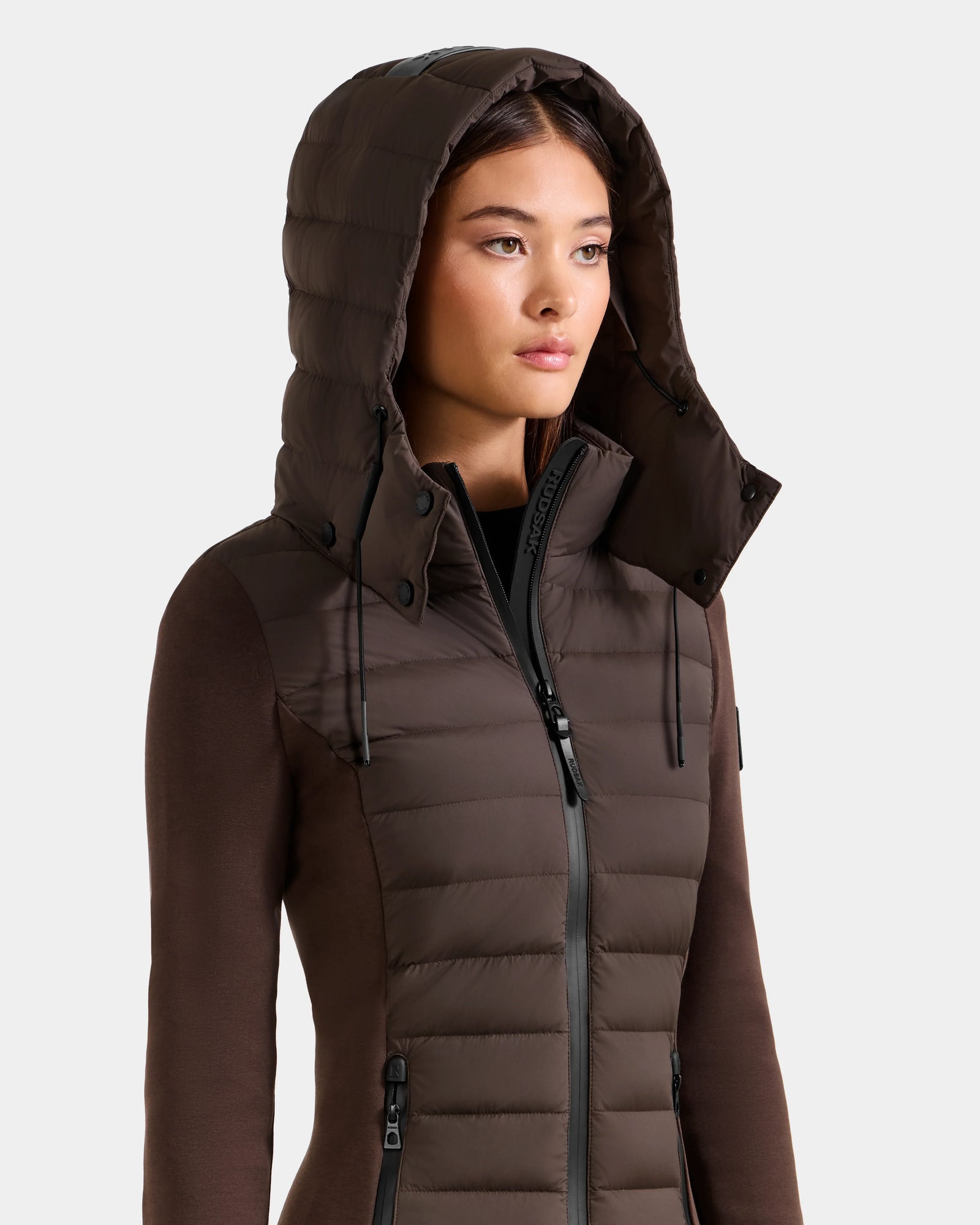 A woman stands against a plain light background, looking to the side, wearing the Rudsak MARRY M - L125519 Espresso women’s hybrid jacket with a high collar and removable hood.