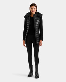 A woman faces forward against a plain light gray background, wearing the Rudsak MARRY S - L125521 Black weather-resistant long puffer vest with a high collar over black attire and heeled boots.