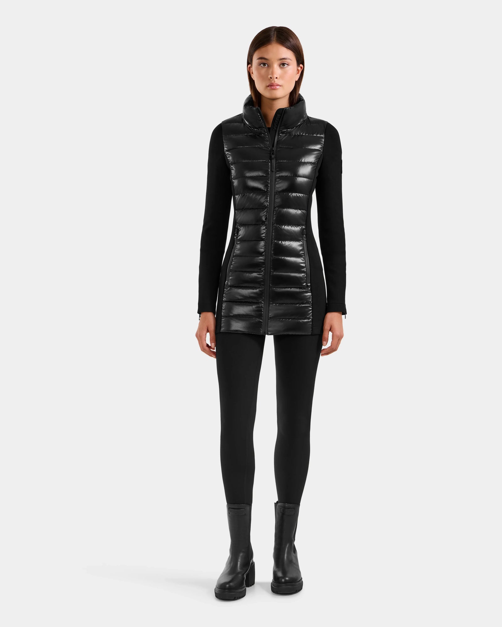 A woman faces forward wearing the Rudsak MARRY S - L125521 Black long, weather-resistant sleeveless puffer vest over a black long-sleeve top, leggings, and ankle boots against a plain light background.