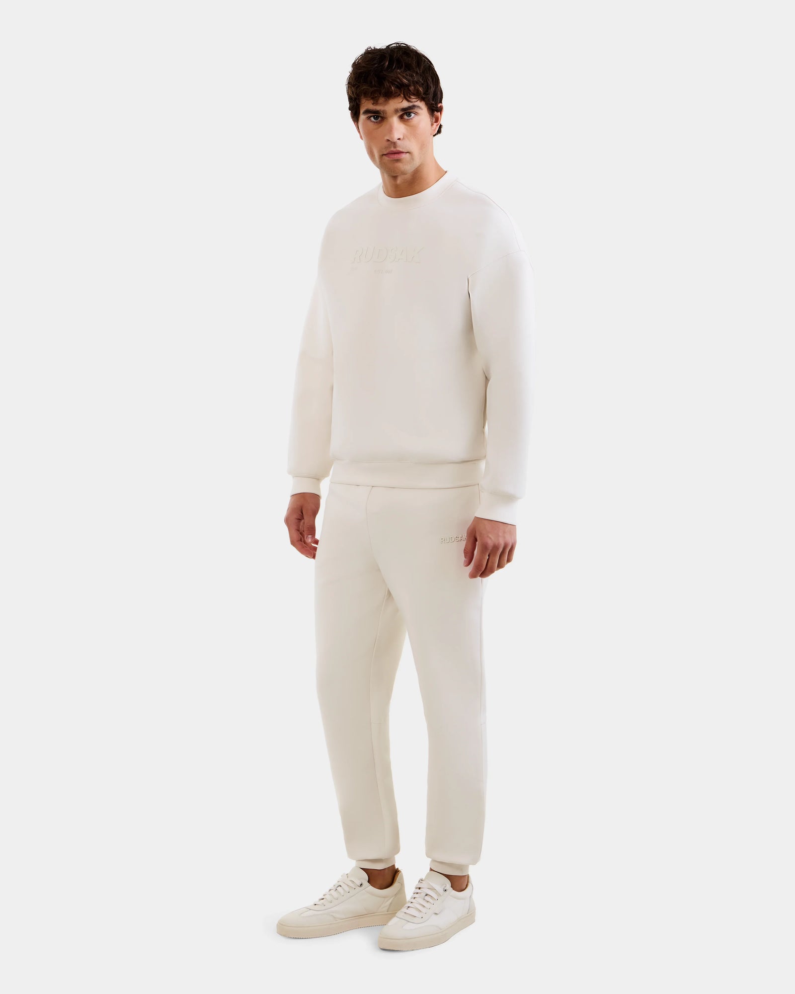 A man faces forward in an all-white outfit, wearing Rudsak's MAXTON - U525007 Vanilla M sweatshirt and unisex jogger pants with sneakers, set against a plain white background.