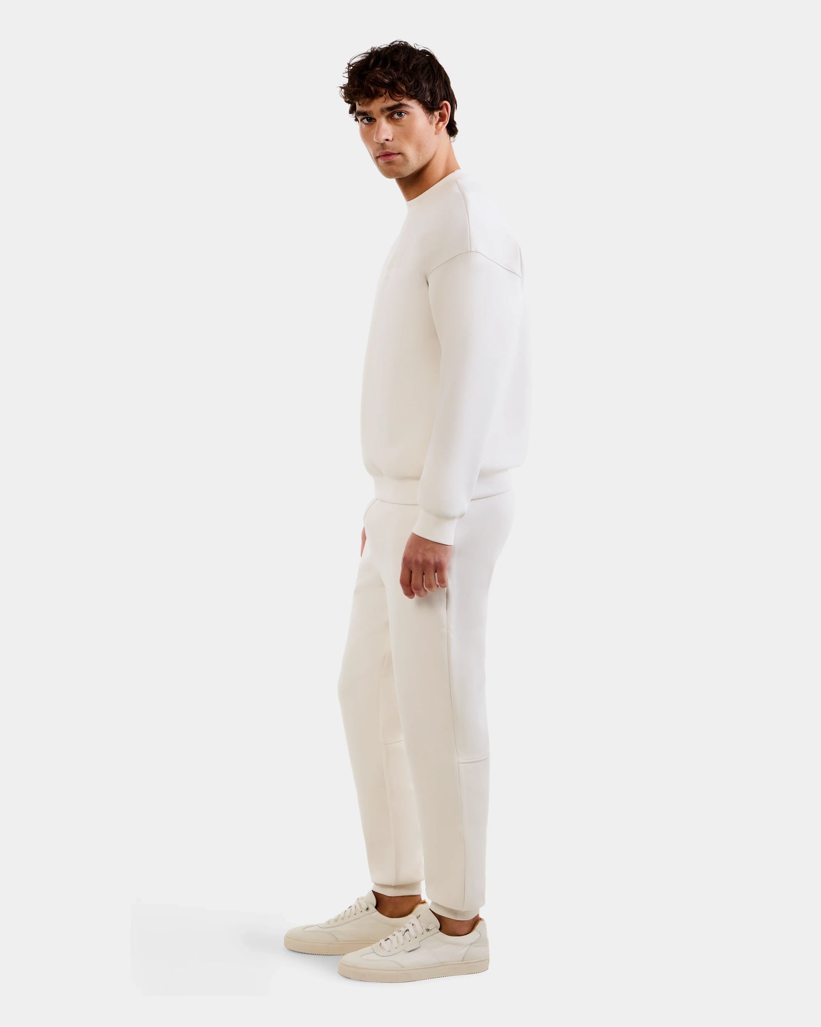 A man stands sideways facing left, wearing the Rudsak MAXTON - U525007 Vanilla M cotton-blend jogger, sweatshirt, and white sneakers against a plain white background for effortless everyday comfort.