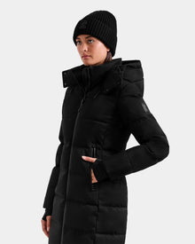 Wearing the Rudsak MIRIAM MW - L125604 Black women’s down puffer coat with a hood and a black knit beanie, she stands against a plain light background, looking slightly to the side with one hand in her pocket.