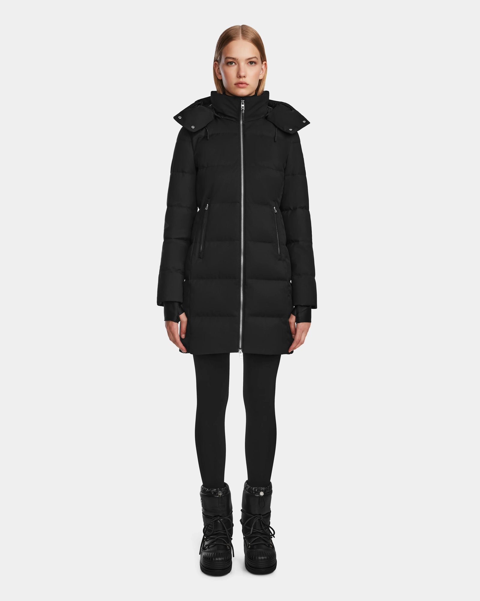 A woman faces forward in the Rudsak MIRIAM - L123544 Black down puffer coat with high collar and hood, paired with black leggings and boots, against a plain white background.