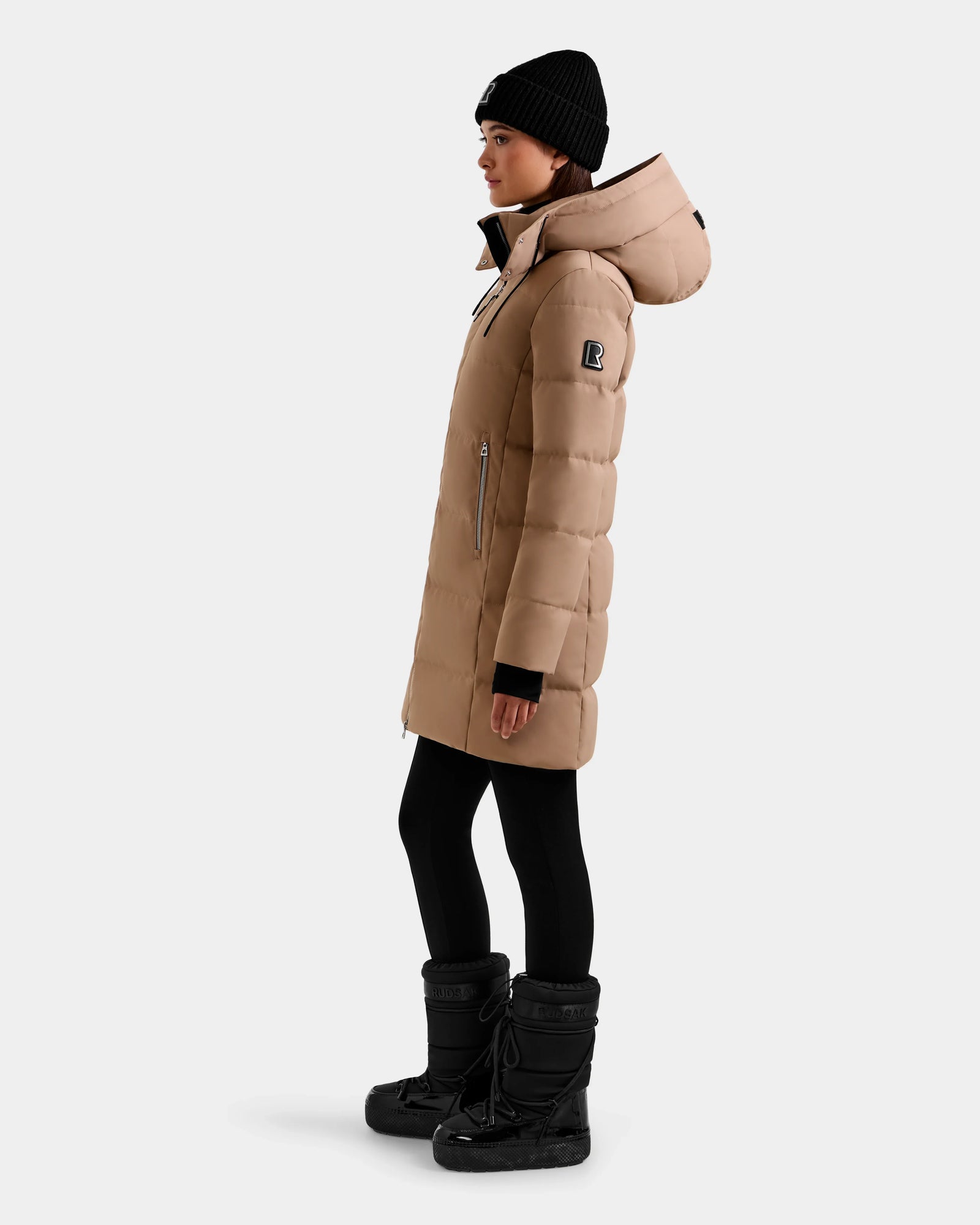 A person stands in profile against a plain light background, wearing the Rudsak MIRIAM - L123544 Sand women's down puffer coat, paired with black leggings, winter boots, and a black knit beanie.