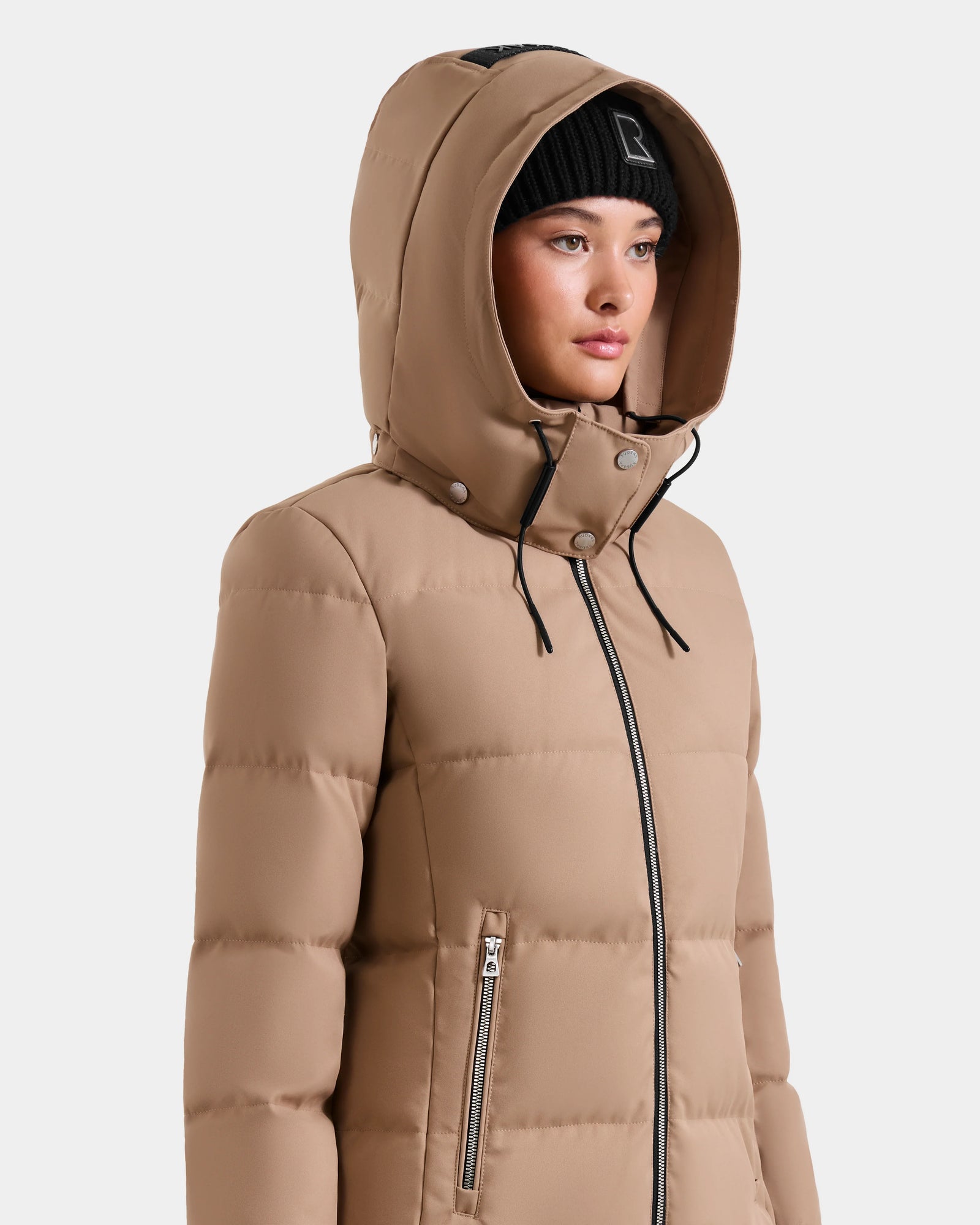 A woman models the Rudsak MIRIAM - L123544 Sand jacket, featuring a weather-resistant tan design with a hood and black zippers, paired with a black knit beanie. She faces slightly right against a white background.