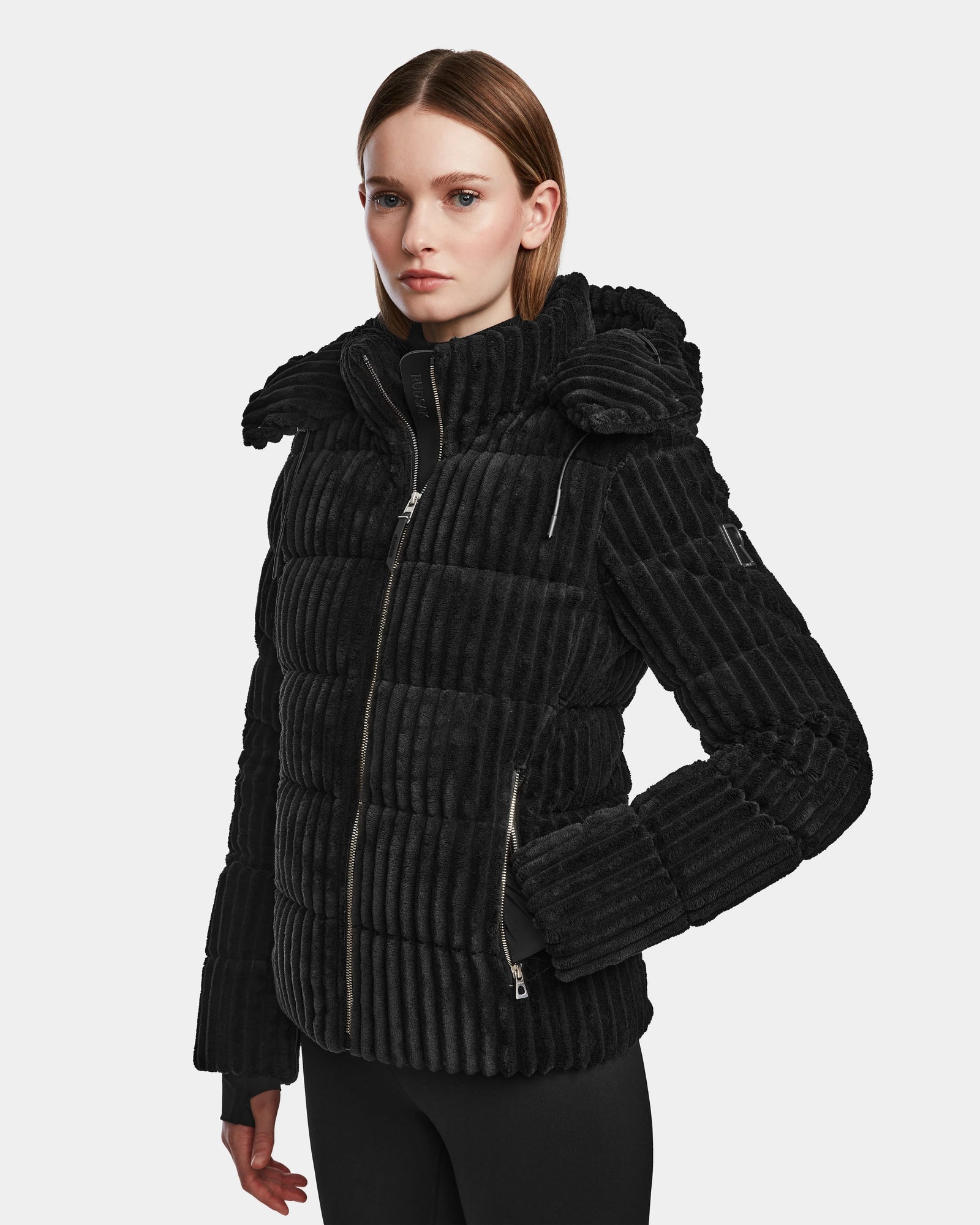 A woman with straight, shoulder-length hair wears the Rudsak MIRRY FF - L124507 Black women's short down puffer jacket, featuring a high collar and zippered pockets, against a plain light gray background.