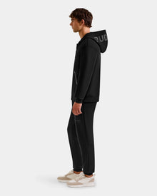 A man stands in profile wearing the Rudsak ROGER - U523507 Black M unisex zip-up hoodie with matching black joggers and white-beige sneakers, set against a plain light gray background.