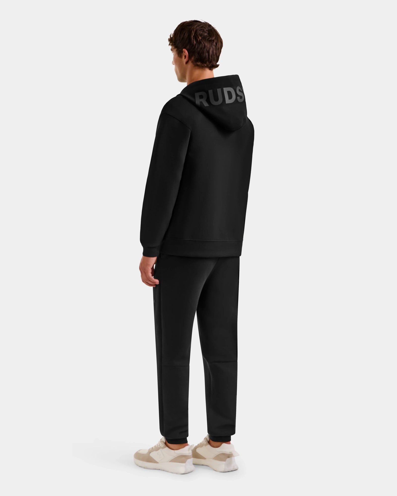 A person faces away, wearing the ROGER - U523507 Black M hoodie by Rudsak, paired with black pants and beige-and-white sneakers—an effortlessly cool urban style set against a plain light background.