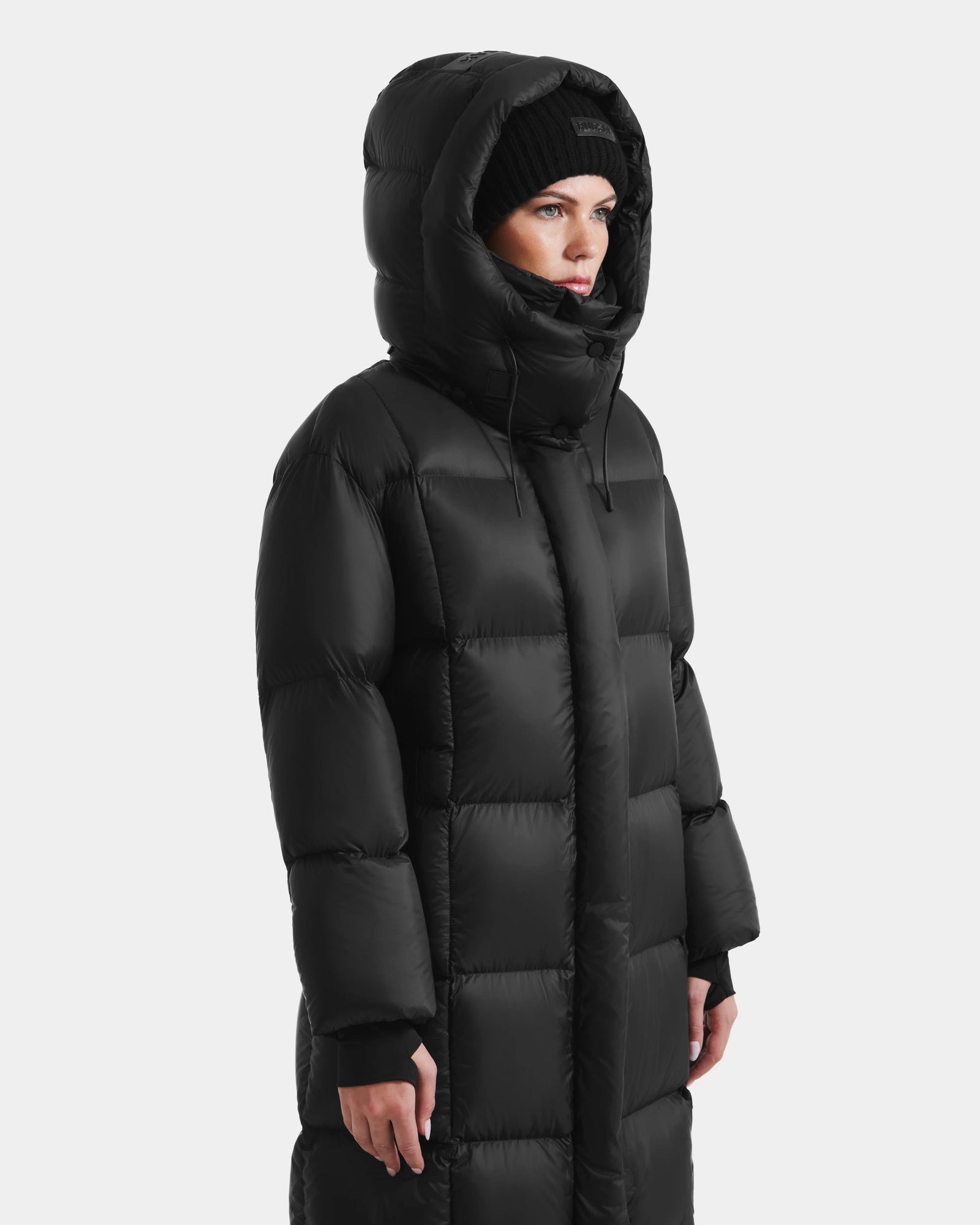 A person wears the Rudsak SASHA - 8125686 Black womens long down puffer coat with removable hood and a black knit beanie, standing against a plain white background.