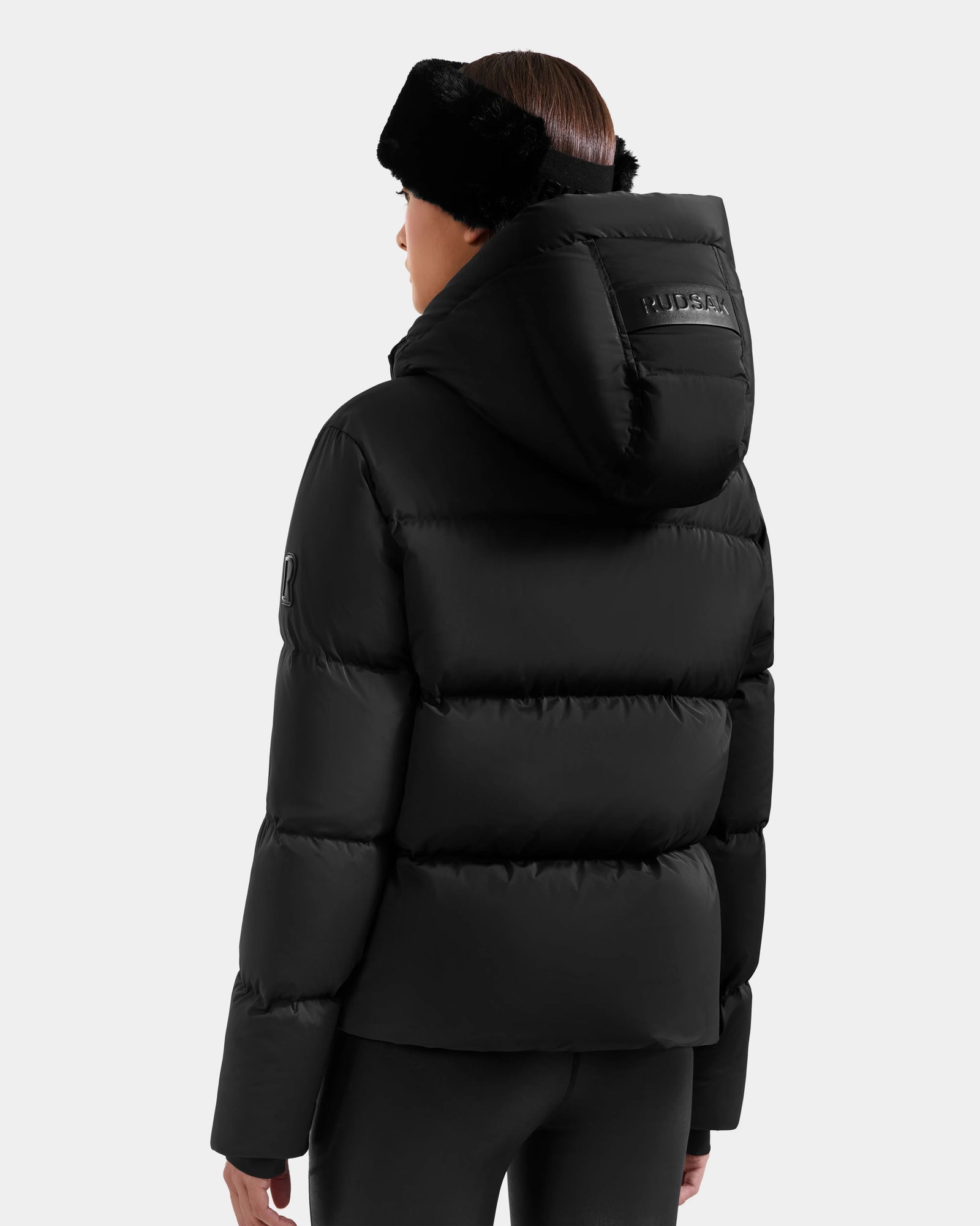 A person is seen from the back wearing the Rudsak STEFANIE M - L125526 Black puffer jacket with a large hood and a black fur headband, set against a plain light gray background.