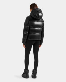 A person with straight brown hair, seen from behind, wears the Rudsak STEFANIE S - L125585 Black shiny puffer jacket with hood, black leggings, and black winter boots against a plain light gray background.