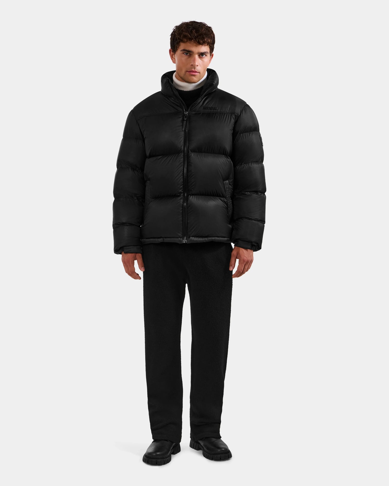 A man stands facing forward against a plain white background, wearing the Rudsak STEFANO - 6125574 Black men’s down puffer winter jacket, paired with black pants and black shoes.