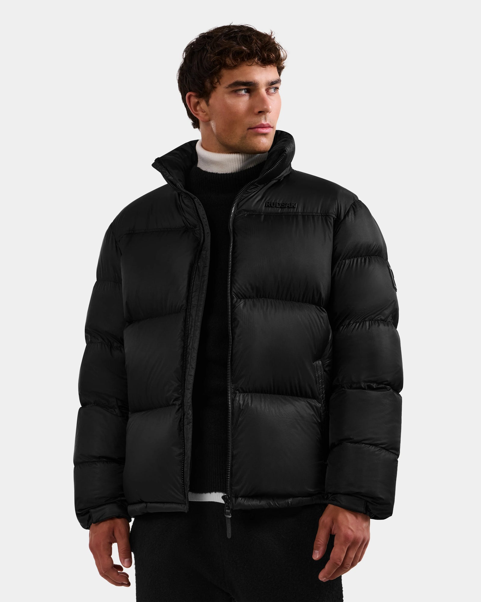 A young man wearing the Rudsak STEFANO - 6125574 Black men’s down puffer jacket over a black sweater and white turtleneck looks to his left against a plain, light background.