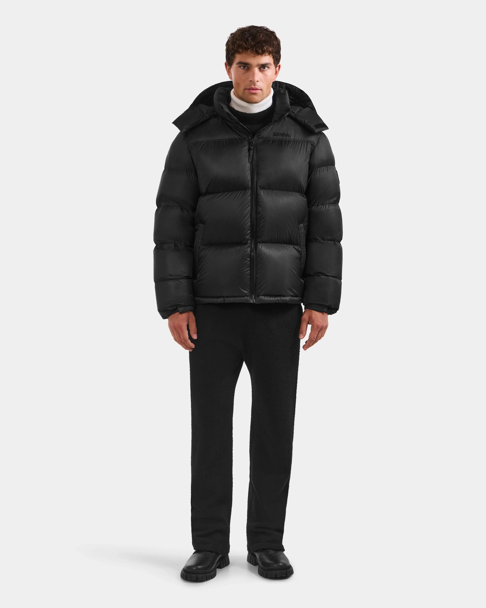 A person stands facing forward in a Rudsak STEFANO - 6125574 Black RDS-certified down jacket with a hood, paired with black pants, black shoes, and a white turtleneck top against a plain light gray background.