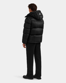 A person faces away, wearing the Rudsak STEFANO - 6125574 Black men’s down puffer jacket with a hood, black pants, and black shoes, set against a plain light gray background.