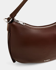 A close-up of the Rudsak SYLVIA - L325515 Mocha women's leather shoulder bag with a crescent silhouette, silver zipper, and short strap, shown against a plain light background.