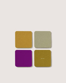 Four square Graf Lantz Merino wool coasters in mustard yellow, sage green, dark gold (with good habits in small gold text), and bright purple are arranged in a 2x2 grid on a light background.