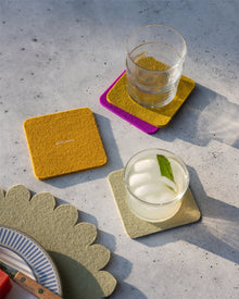 Two drinks in clear glasses rest on vibrant Graf Lantz Merino wool coasters atop a gray surface—one with ice and mint, one empty. A scalloped placemat and plate appear in the lower left corner.