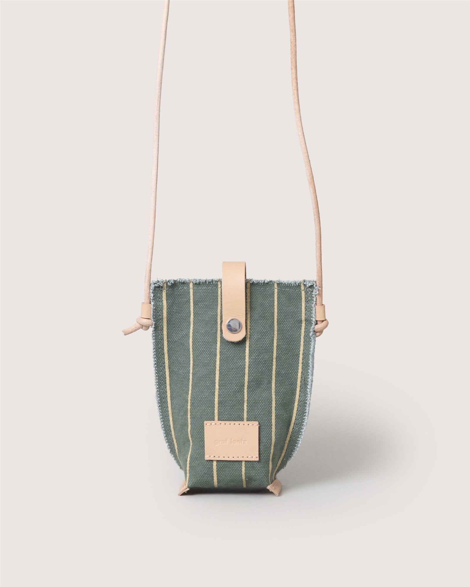 A small green Graf Lantz Hana Canvas Phone Crossbody features vertical yellow stripes, a beige leather snap closure, matching strap, and a rectangular beige leather logo patch on the front, shown against a light background.
