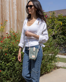 A woman wearing sunglasses, a white shirt over a tank top, and blue jeans strolls outdoors by green bushes and a wooden wall, carrying a compact Graf Lantz crossbody bag for a stylish hands-free look.