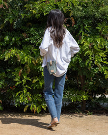 A person with long dark hair, dressed in a white oversized shirt, blue jeans, and brown shoes, carries a compact Graf Lantz crossbody bag while standing on a dirt path facing leafy green bushes.