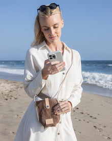 A woman in a white dress stands on a sandy beach near the ocean, smiling at her smartphone. Sunglasses rest on her head, and she carries the Graf Lantz Hana Canvas Phone Crossbody for a stylish, hands-free look.