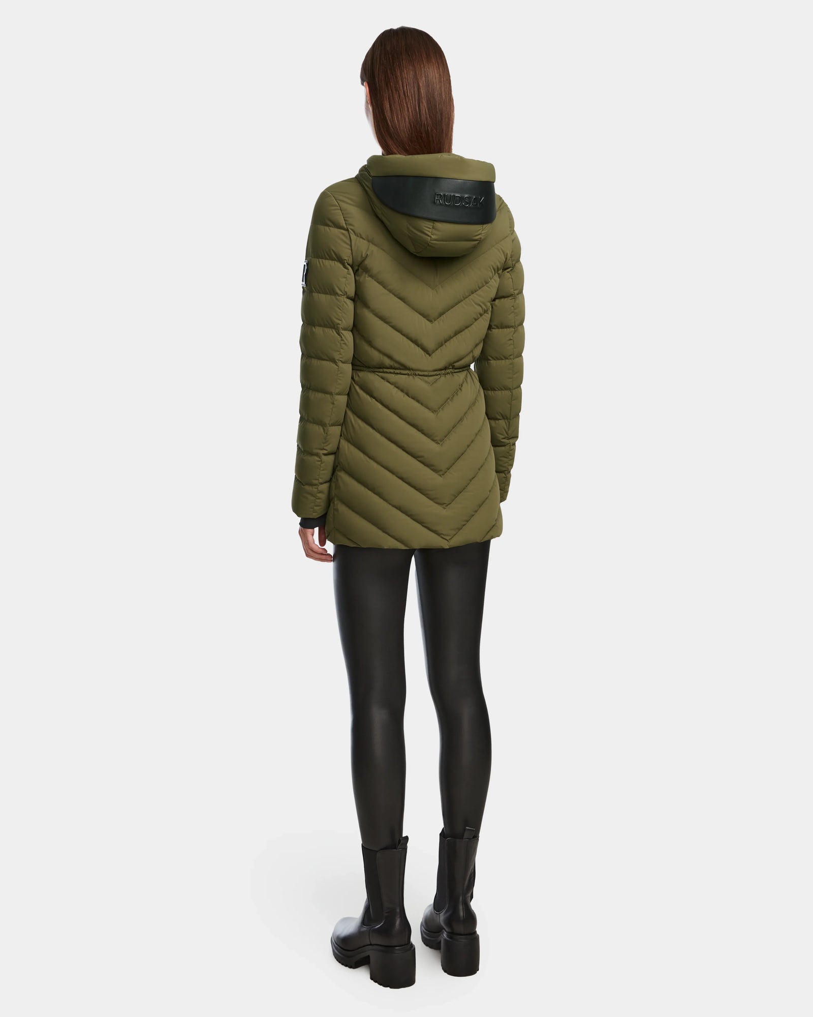 A person faces away, wearing the Rudsak FRANCINE - 8124722 Loden Green quilted puffer hooded jacket with RDS-certified down, black leggings, and black boots against a plain white background.