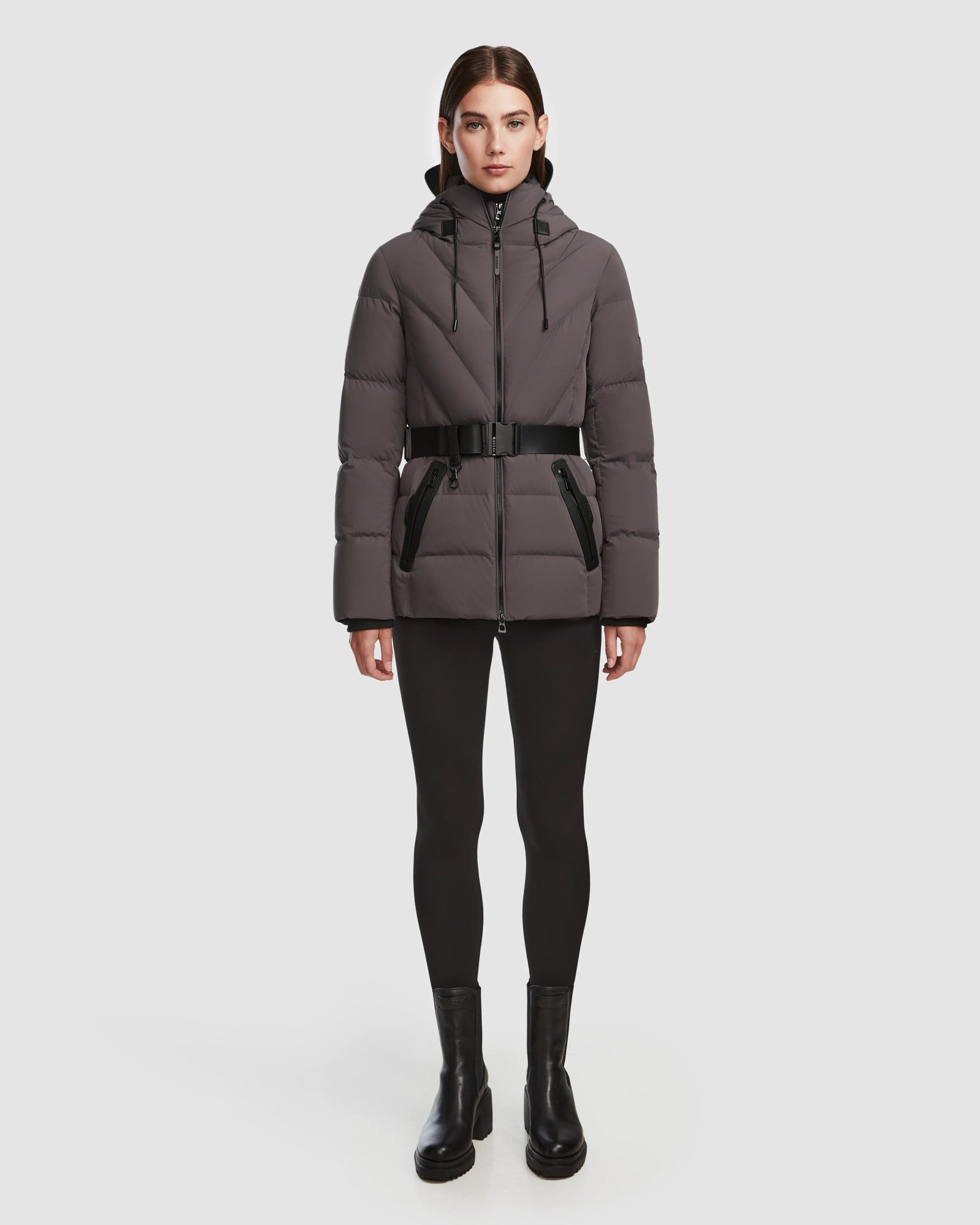 A person stands facing forward in a Rudsak ENZAH - 8124692 Grey down jacket with a black belt, paired with black leggings and ankle boots against a plain white background.