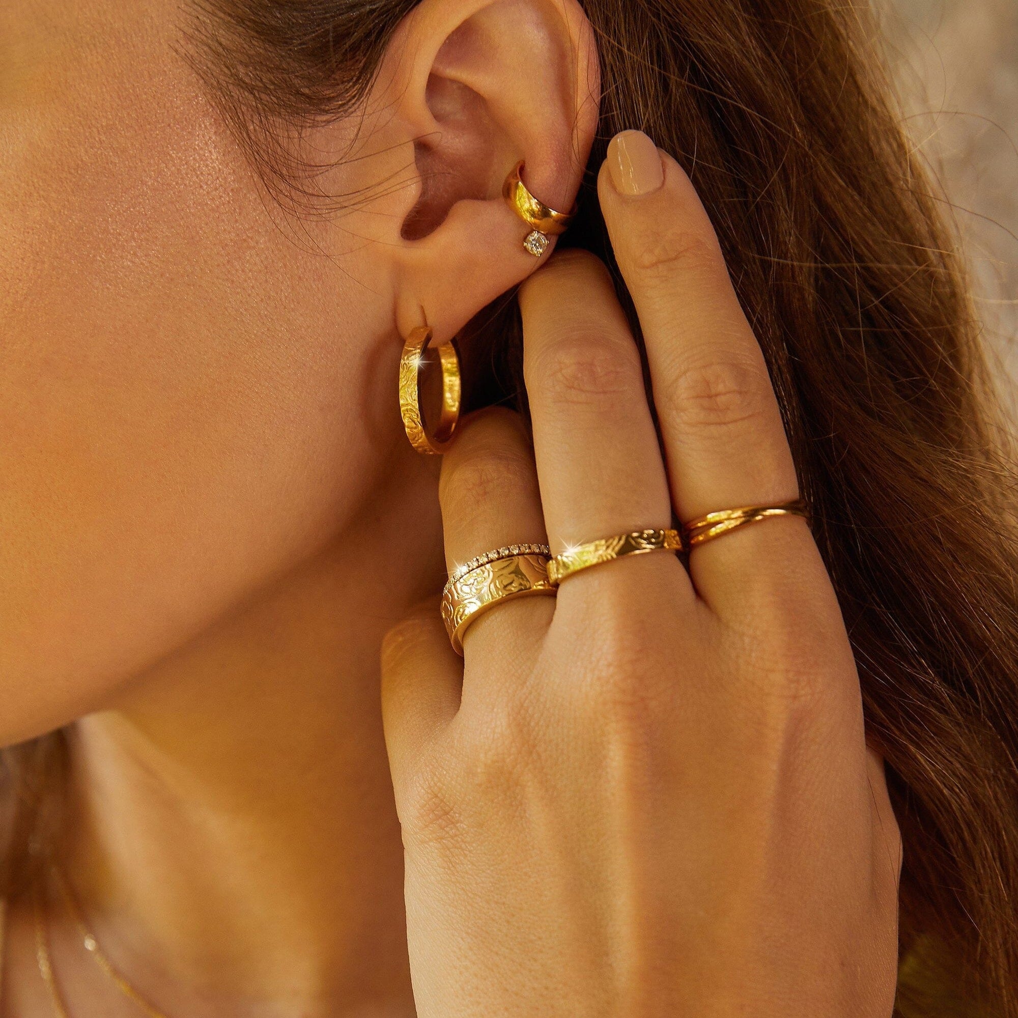 Lifestyle Model Close-up of a woman resting her hand near her ear, wearing gold hoop earrings and multiple gold rings.