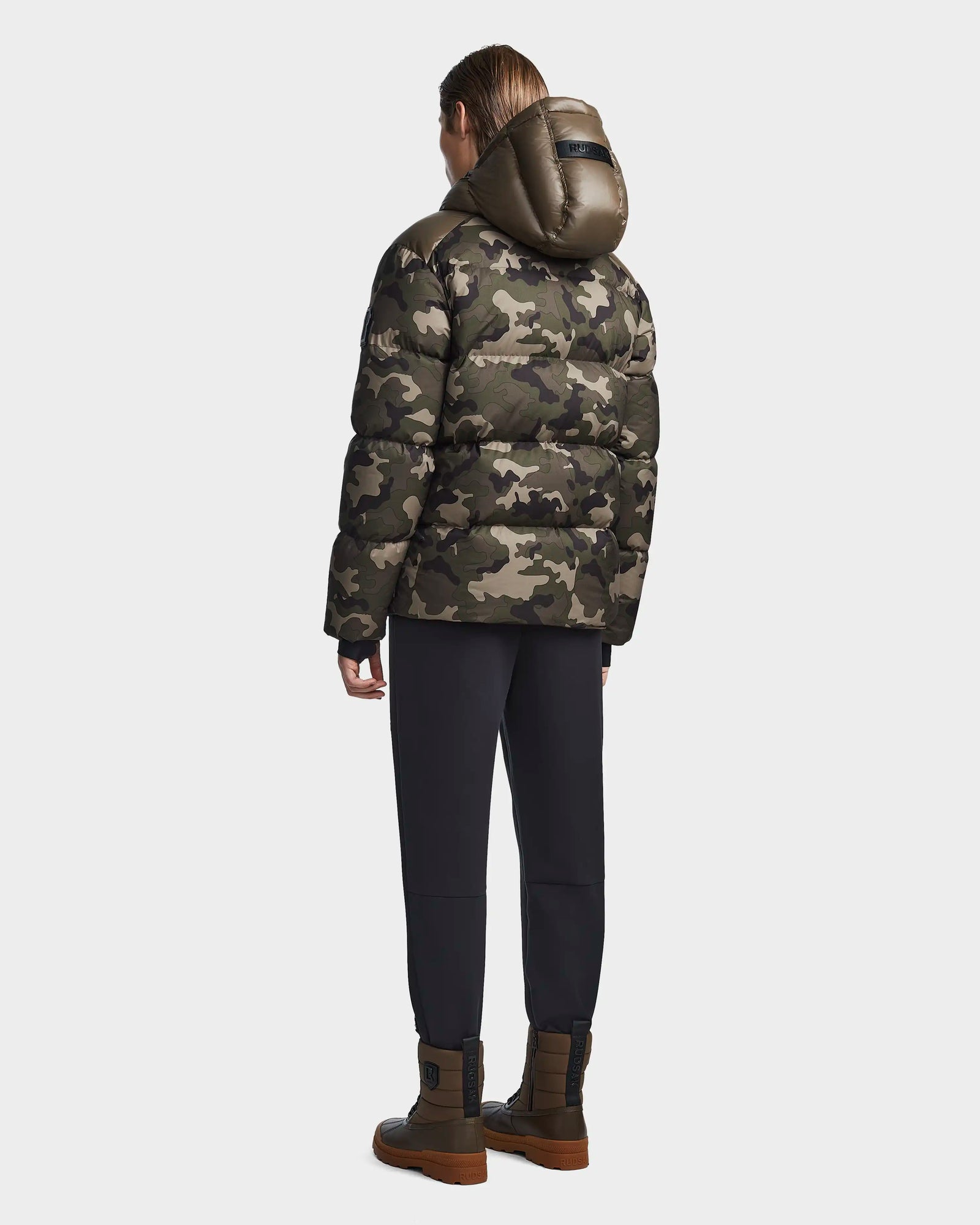 A person stands facing away, wearing the Rudsak RONNY M - M123512 Camouflage mens weather-resistant jacket with a hood, paired with dark pants and brown high-top boots, set against a plain light background.