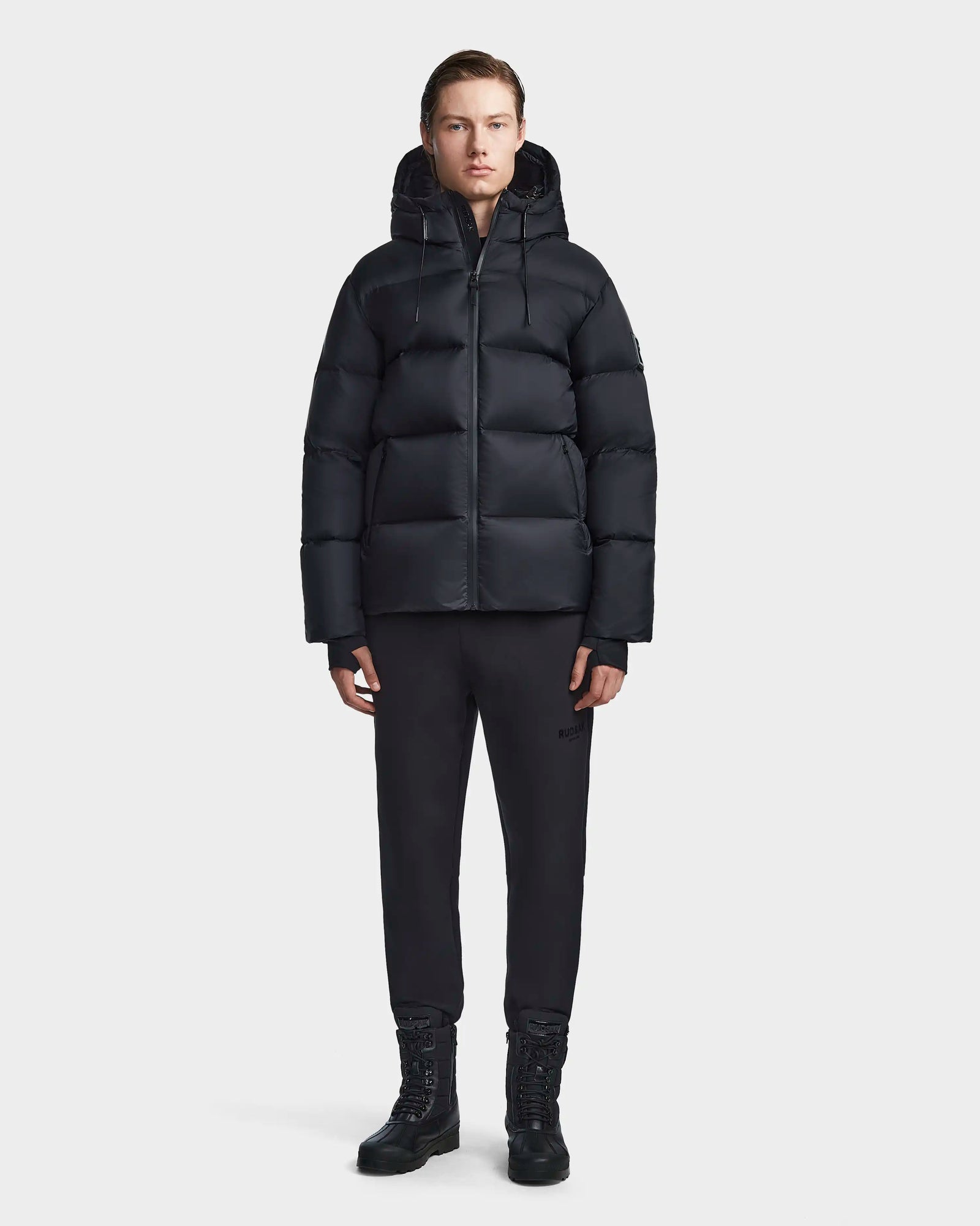 A person with short hair stands against a plain background, facing forward with arms relaxed, wearing the Rudsak DANIEL M - M123510 BLACK mens coat with a hood, paired with black pants and black boots.