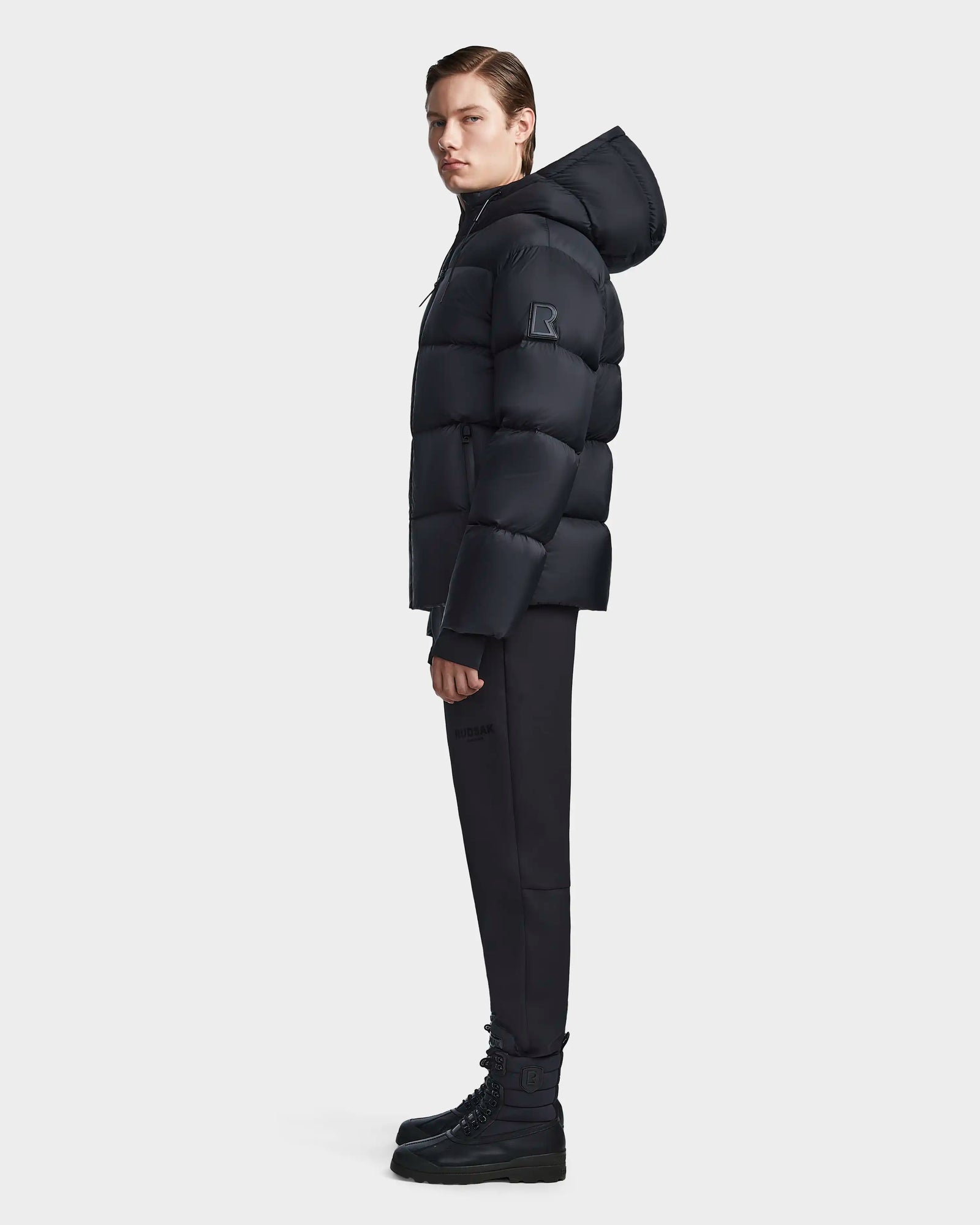 A person stands sideways against a plain light background wearing the Rudsak DANIEL M - M123510 BLACK weather-resistant winter jacket, matching black pants, and black boots.