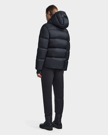 A person faces away from the camera, wearing the Rudsak DANIEL M - M123510 BLACK mens hooded down puffer jacket, styled with black pants and boots against a plain light backdrop.